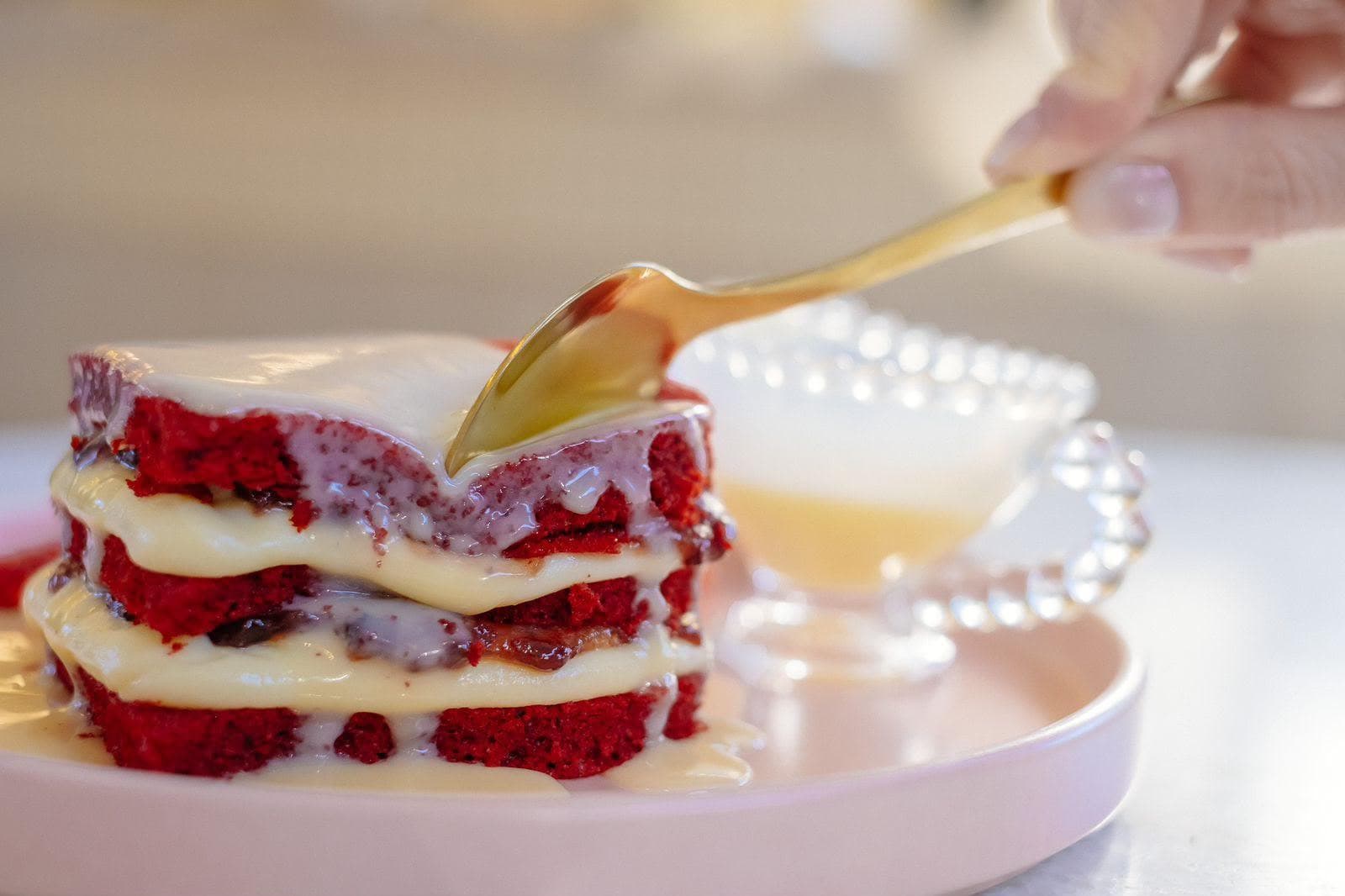 Bolo red velvet em três camadas, com creme branco entre elas, recebendo calda servida de uma colher dourada sobre um prato rosa
