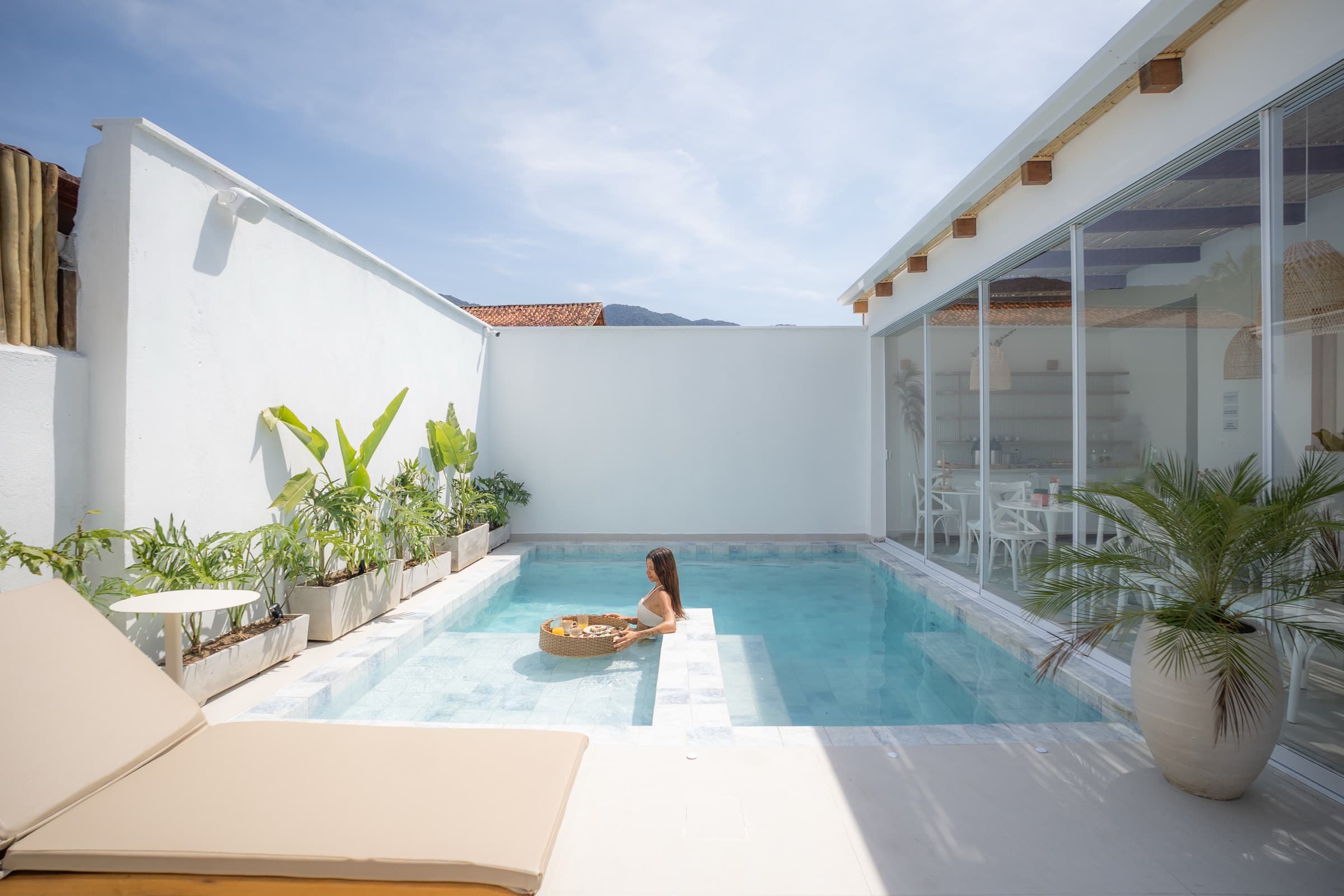 Pátio da piscina entre muros brancos caiados: uma hóspede na água ao lado de uma bandeja flutuante de vime, o salão envidraçado à direita e a serra atrás do telhado