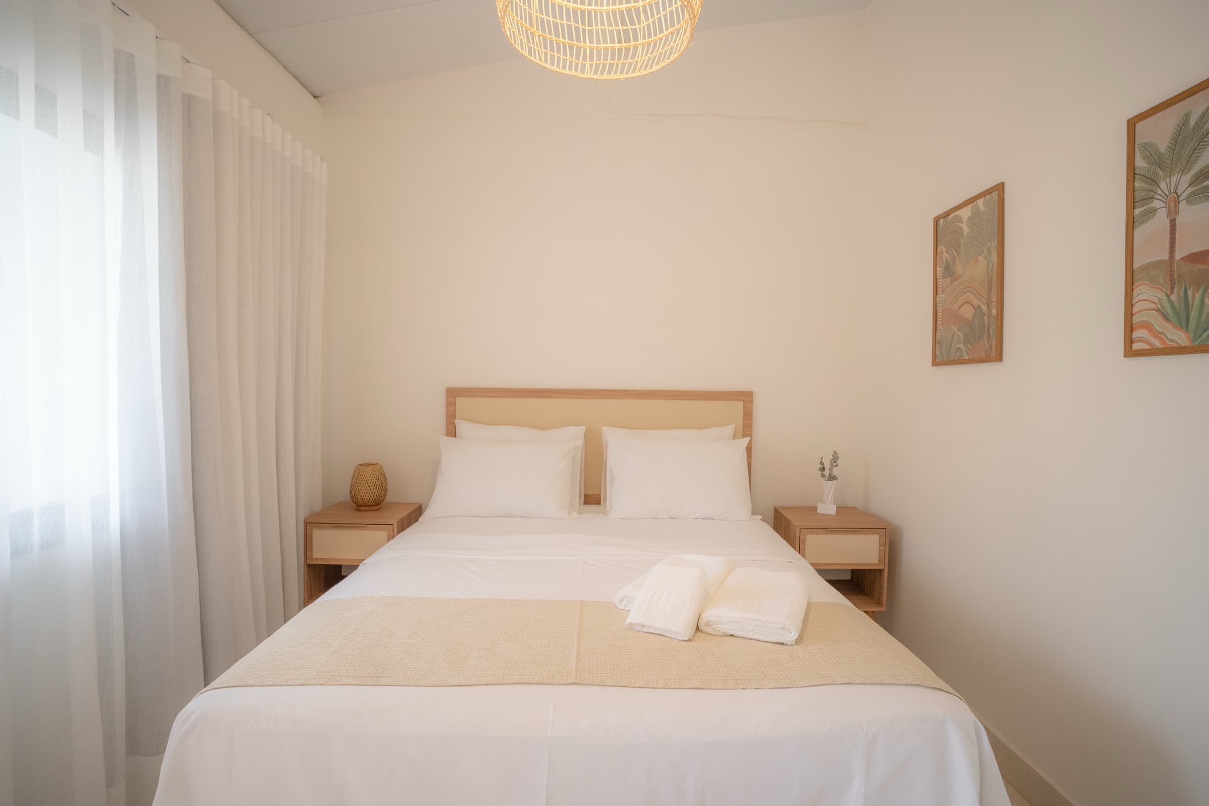Cama de casal com cabeceira de madeira clara e roupa de cama branca, pendente de fibra trançada, dois quadros botânicos e cortina de voil na janela